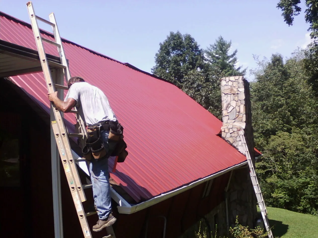 Roofer on a ladder beside a red metal roof and stone chimney — Soffit & Fascia Repair by Roofing Brothers in Lapel